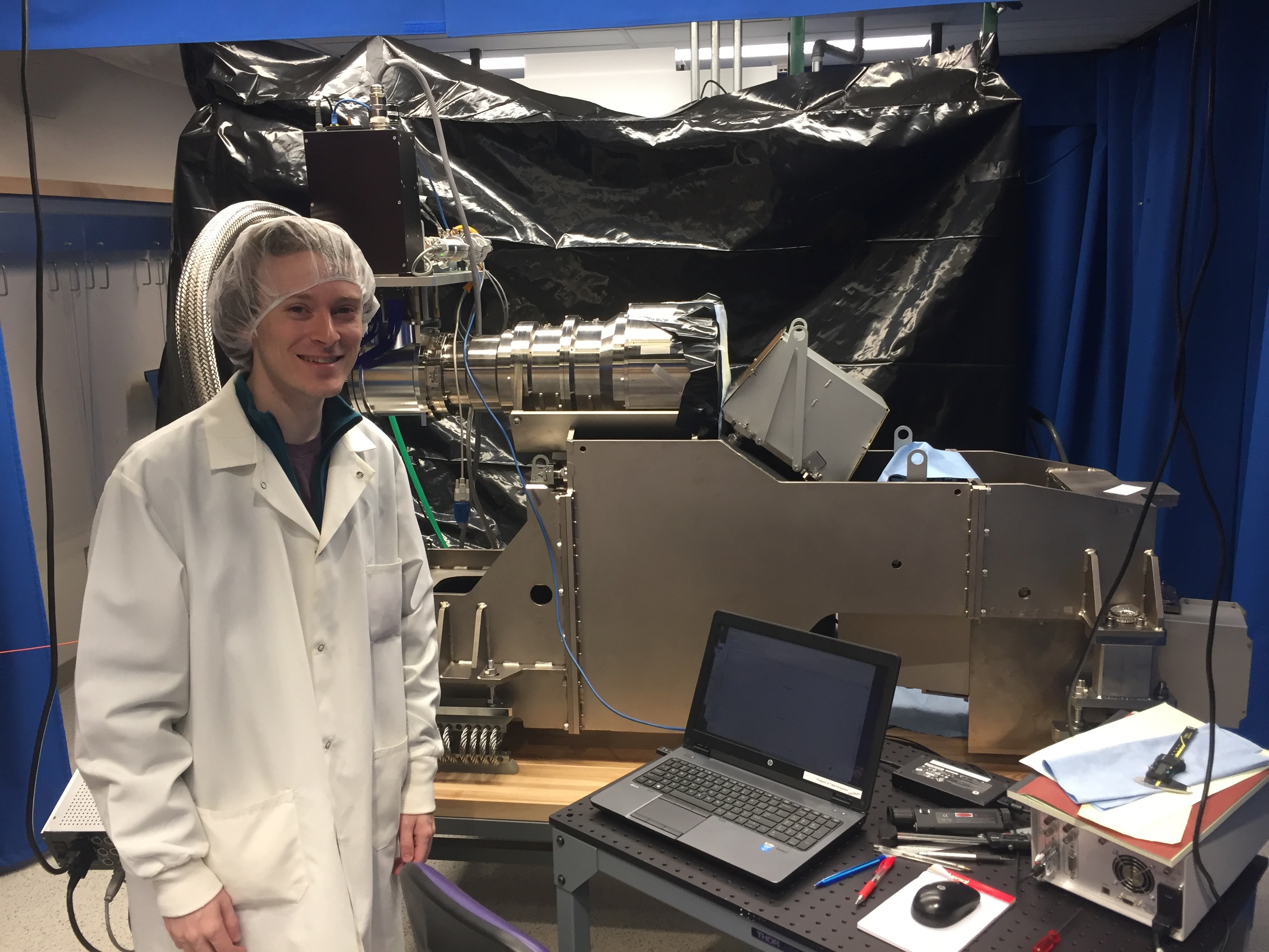 Allen in a lab coat and hair net standing next to the EXPRES spectrograph during construction at Yale.